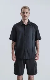 Camisa de manga corta en color negro con proceso de lavado, cuello clásico y cierre frontal con botones. Presenta un diseño minimalista con una costura vertical decorativa en el frente.