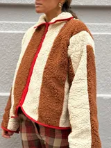 Campera de corderito con diseño de rayas verticales en colores marrón y crema, con cierre frontal y detalles de ribete en color rojo.