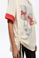 Remera larga de color marfil con detalle de abertura y lazos en el hombro, estampa frontal de zapatillas de ballet con lazos rojos y mangas cortas con puños en contraste rojo y beige. Cuenta con cordón ajustable lateral.