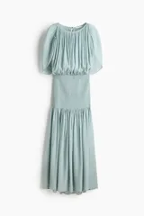 Vestido midi de tejido texturizado color verde menta, con escote redondo fruncido, espalda semiabierta con breteles finos y capa decorativa sobre los hombros. Cuenta con sección de punto smock en la cadera y falda con leve vuelo.