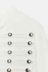 Chaqueta de denim blanca con cuello redondo, manga larga y corte entallado. Presenta un diseño de inspiración militar con múltiples tiras horizontales en el delantero decoradas con botones metálicos plateados, y cierre frontal con ganchos.