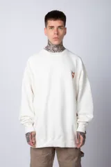 Buzo de tejido friza color crema, de corte oversize y cuello redondo. Presenta un pequeño bordado o parche en el pecho izquierdo con un diseño de cabeza de animal en tonos rojizos/terracota.