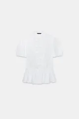 Camisa blanca de manga corta abullonada, con cuello mao y cierre frontal con alamares. Presenta corte evasé con detalle de volados en el bajo.