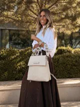 Mochila tipo cartera con tapa, confeccionada en símil cuero color suela. Cuenta con doble compartimento, división interna, cierre de seguridad y bolsillo trasero con cierre. Incluye una correa frontal decorativa con tachas metálicas.