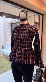 Blusa de manga larga con estampado a cuadros en tonos beige y negro, cuello alto con volados, diseño fruncido en el pecho y las mangas, y terminación acampanada en los puños y el bajo.
