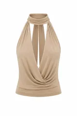 Top de tela alicrada color beige, con cuello halter y diseño de caída tipo buche en el frente. Presenta espalda descubierta y cierre anudado al cuello.
