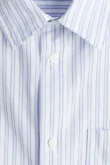 Camisa de manga corta en popelina de algodón a rayas verticales celestes y blancas. Modelo boxy con cuello inglés, tapeta clásica, bolsillo superior abierto y canesú en la espalda. Bajo recto con aberturas laterales.