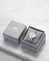 Reloj de pulsera analógico con caja redonda de metal plateado y malla de acero tipo milanesa. Presenta una esfera minimalista con números romanos y manecillas finas.