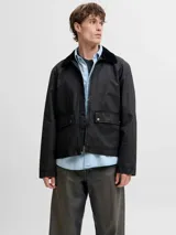 Campera de estilo worker en color negro, con acabado recubierto, cuello camisero en contraste, cierre frontal y bolsillos con solapa y broches a presión.