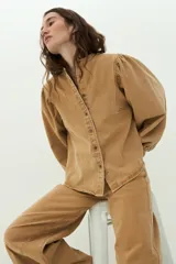 Camisa de jean color camel, de manga larga abullonada, con cuello mao y cierre frontal de botones metálicos.