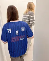 Camiseta de fútbol de manga corta en color azul con diseño estampado abstracto y cuello en V rosado con detalles blancos a juego con los puños. Presenta inscripciones y escudos deportivos en el pecho.