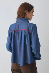 Camisa de denim azul con cuello alto tipo gorguera, cierre frontal con botones de madera y detalle de volados laterales. Presenta mangas largas con puños elásticos terminados en volados y detalles de costuras en color rojo en hombros y puños.
