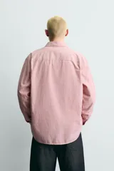 Camisa de corte relajado confeccionada en tejido de algodón, con diseño de rayas verticales en tonos rosados y blancos. Presenta cuello abotonado, cierre frontal de botones, manga larga con puños abotonados y bajo asimétrico.