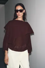 Jersey de punto color vino tinto, de corte amplio y holgado, con cuello bote y un detalle de capa asimétrica superpuesta en la parte superior delantera. Mangas anchas tipo murciélago.