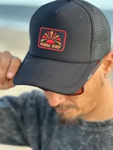 Gorra trucker negra con paneles frontales rígidos y paneles traseros de malla. Presenta un parche bordado en el frente con un diseño abstracto en tonos naranja, amarillo y blanco.