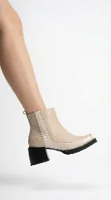 Bota corta de cuero en tono beige, con diseño bicolor que presenta un panel lateral en un tono más claro. Cuenta con un taco cuadrado negro de altura media y cierre lateral interno.