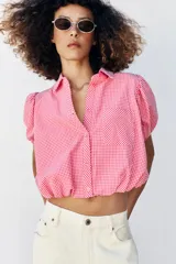 Camisa corta de cuello solapa y manga corta abullonada acabada en elástico, color verde con estampado de cuadros vichy. Cuenta con un bolsillo de plastrón en el delantero, bajo con elástico y cierre frontal con botones.