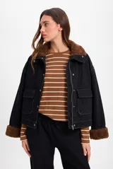 Chaqueta de largo medio y silueta holgada, confeccionada en tela de jean color negro con costuras a contratono. Presenta forro interno de corderito color marrón, cuello alto, cierre frontal con cremallera y botones a presión, y bolsillos plaqué con tapa en el frente.
