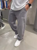 Pantalón deportivo de corte baggy y ajuste holgado, color gris oscuro. Presenta costuras verticales frontales que recorren las piernas y un pequeño logo bordado en la parte superior. Cuenta con bolsillos laterales y cintura elástica.