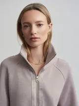 Campera deportiva con cierre frontal, cuello alto tipo chimenea y mangas raglán. Es de corte holgado y presenta bolsillos de parche delanteros. El color es un tono neutro entre gris y rosado (taupe).