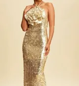 Vestido largo de fiesta totalmente cubierto de lentejuelas doradas, con escote halter y un detalle de flor grande tridimensional en el pecho. Presenta una silueta ajustada que se extiende hasta el suelo.