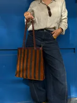 Cartera tipo tote de cuero con diseño de franjas verticales multicolores en tonos tierra, rojo y azul. Cuenta con correas trenzadas de cuero y cierre superior.