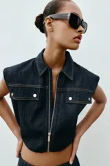 Chaleco de denim oscuro con cuello solapa, cierre frontal con cremallera metálica y bolsillos delanteros con tapeta. Presenta un diseño de corte abullonado con bajo elástico.