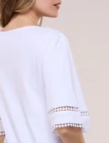 Remera blanca de silueta recta y algodón suave, con escote redondo y pequeña abertura central tipo gota. Presenta mangas de largo intermedio con aplicación horizontal de puntilla calada al tono y ruedo recto con tajos laterales.