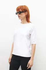 Remera blanca de manga 3/4 con cuello redondo y botones metálicos decorativos en los hombros.