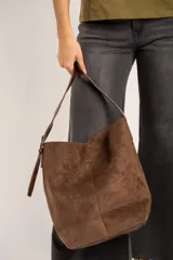 Cartera tipo shopper de gamuza sintética color marrón, con diseño estructurado, asa de hombro ajustable y detalle de placa metálica dorada con logo en la parte frontal.