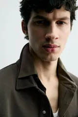 Sobrecamisa de corte regular fit en tejido elástico y transpirable, con cuello solapa, cierre frontal mediante botones a presión y bolsillo de plastrón en el pecho. Presenta mangas largas con puños abotonados y bajo con aberturas laterales.