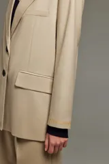 Blazer color beige de lana, con calce recto, solapas de muesca, cierre frontal con dos botones metálicos personalizados, bolsillos con solapa y bordado dorado en la manga.