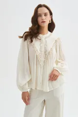 Blusa de algodón color beige, con cuello redondo y cierre frontal con botones. Presenta delicados bordados decorativos en tono tostado sobre el pecho y los hombros, mangas largas abullonadas con puños ajustados y diseño holgado.