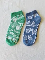 Pack de dos pares de soquetes tobilleros estampados. Un par es verde con estampado de hojas y flores blancas, y el otro es azul con estampado de mariposas blancas.