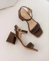 Sandalias de gamuza color marrón chocolate, de taco cuadrado medio y punta cuadrada. Tienen una tira delantera ancha y una tira ajustable al tobillo con hebilla metálica dorada.
