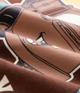 Pijama corto de algodón compuesto por una remera marrón de manga corta con cuello redondo y un pequeño estampado ovalado con texto en el pecho, y una bermuda con diseño a cuadros en tonos marrones y beige.