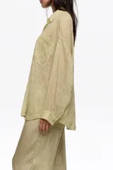 Camisa de manga larga color beige con textura plisada, cuello camisero, cierre frontal de botones, bolsillo en el pecho y bajo redondeado.