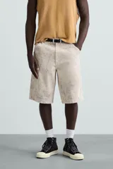 Bermuda de algodón color beige claro con efecto lavado y estampado abstracto en tonos grises. Diseño de corte relajado con cierre frontal de botón y cremallera, bolsillos delanteros y bolsillos traseros de plastrón.