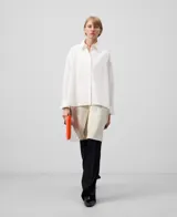 Camisa oversize blanca de manga larga con cuello solapa y botonadura frontal. Presenta una pieza superpuesta en la espalda que corresponde al delantero de un blazer.