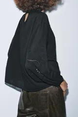 Camisa negra de manga larga con cuello redondo. Presenta detalles de entredós y jaretas verticales en el delantero, y cierre de botones en la espalda.