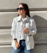 Camisa de cuero ovino italiano color blanco, de corte holgado, con cuello clásico, cierre frontal con broches metálicos, bolsillos laterales y puños con broches.