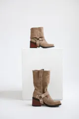 Bota de caña media confeccionada en cuero gamuzado color chocolate. Presenta un diseño de estilo western con taco cuadrado de 7 cm y una correa decorativa en el tobillo con herrajes metálicos plateados.