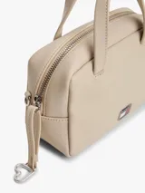 Cartera de mano tipo bowling de Tommy Jeans, color beige, con doble asa superior y correa ajustable extraíble. Presenta un diseño estructurado con cierre superior y logo metálico de la marca en la parte frontal.