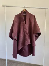 Ruana de estructura rígida confeccionada en tela tipo tapiz, con diseño abierto y caída asimétrica. Presenta un tono magenta con textura jaspeada y etiqueta de marca visible en el frente.