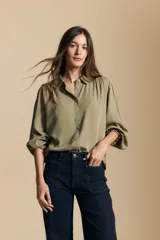 Camisa de manga larga color verde oliva, con cuello clásico, cierre frontal de botones y puños elásticos con detalle de volados. Presenta un diseño de calce fluido con frunces en los hombros y espalda.
