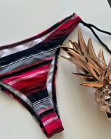 Conjunto de bikini con estampado de rayas horizontales en tonos fucsia, rojo, negro y gris. El corpiño es de estilo triangular con tirantes finos que se atan al cuello y la espalda. La bombacha es de corte colaless con tiras laterales ajustables.