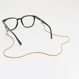 Correa para lentes dorada con cadena de eslabones trenzados y terminales de goma transparente ajustables. Incluye dije circular con el nombre de la marca grabado.