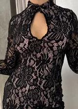 Vestido corto de encaje negro con forro en tono nude, de corte ajustado, cuello alto y abertura frontal tipo gota. Presenta mangas largas con terminación acampanada.