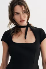 Vestido corto de color negro con escote cuadrado y detalle de lazada en el cuello. Presenta mangas cortas y cierre de cremallera oculta en la espalda.