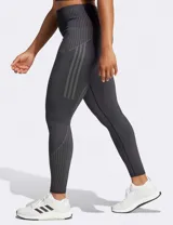 Calza deportiva Adidas Seamless 7/8 de tiro alto, color negro, confeccionada con tejido suave y elástico sin costuras con tecnología AEROREADY. Presenta diseño de líneas acanaladas en la cintura y los tobillos, y las tres tiras características de la marca en el muslo derecho.